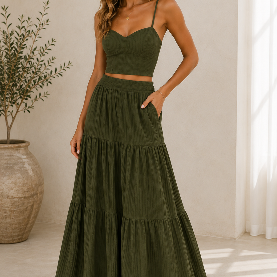 A woman modeling a matching two-piece set consisting of an olive green spaghetti-strap crop top and a high-waisted, tiered maxi skirt with side pockets, set against a warm beige wall with a potted plant.