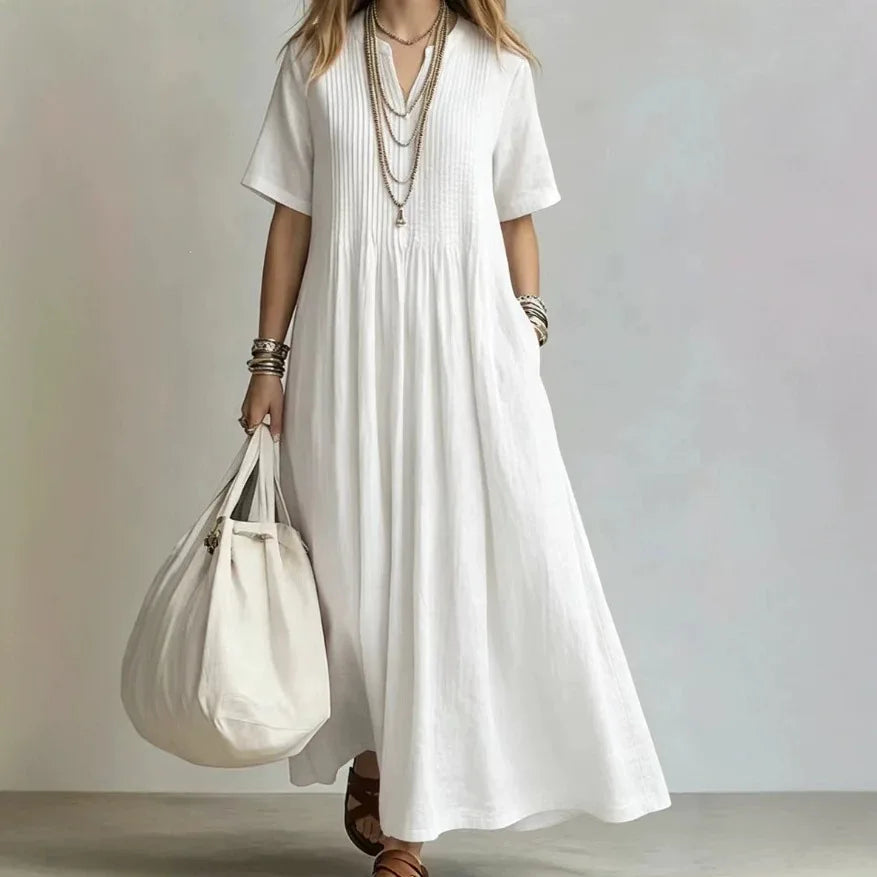 A woman wearing a loose white short-sleeve maxi dress with pleated detailing, holding a large cream canvas tote bag. She is accessorized with layered gold necklaces and bracelets, standing against a neutral light gray background.