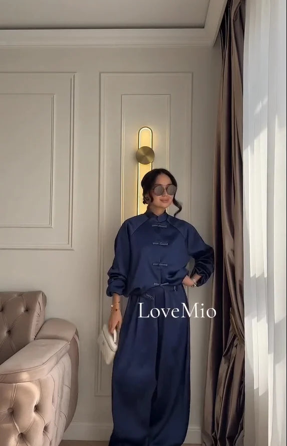 A woman wearing a navy blue traditional Chinese-style long-sleeve top and matching wide-leg pants stands in a decorated room with a tufted sofa, wall sconce, and curtains. The brand name 'LoveMio' is overlaid on the image.