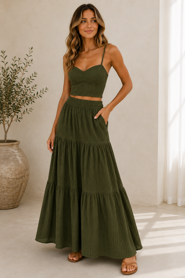 A woman modeling a matching two-piece set consisting of an olive green spaghetti-strap crop top and a high-waisted, tiered maxi skirt with side pockets, set against a warm beige wall with a potted plant.