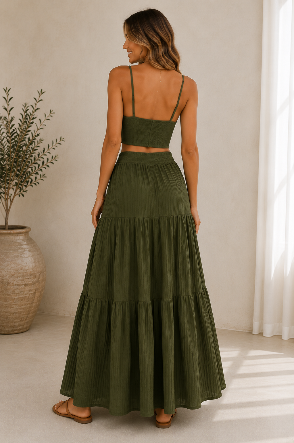 A woman shown from the back wearing an olive green two-piece set consisting of a cropped spaghetti-strap top and a tiered maxi skirt, standing in a brightly lit room with a potted plant on the left.