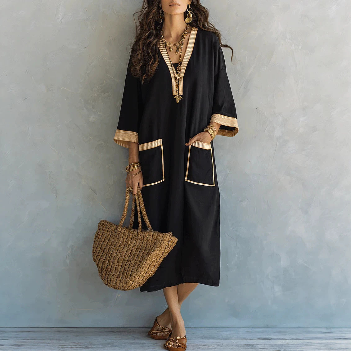 A woman models a black kaftan dress with beige trim, holding a woven straw tote bag against a textured gray wall.