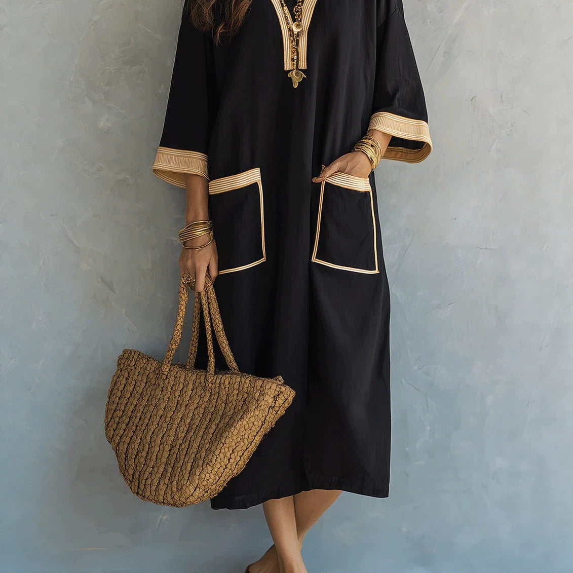 A woman wearing a black kaftan dress with beige trim and pocket details, holding a woven straw tote bag against a textured blue-gray wall.