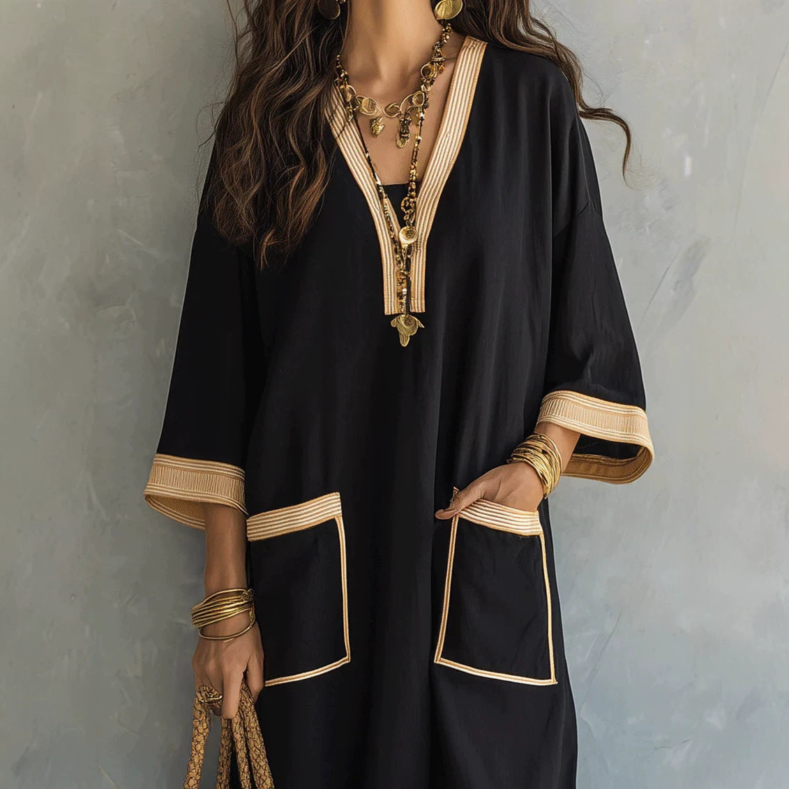 A woman wearing a loose black kaftan-style tunic with beige striped trim along the V-neck, cuffs, and pocket edges, accessorized with layered gold necklaces and bracelets against a textured gray wall.