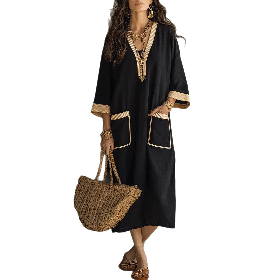 A woman models a black kaftan dress with beige trim, holding a woven straw tote bag against a plain white background.