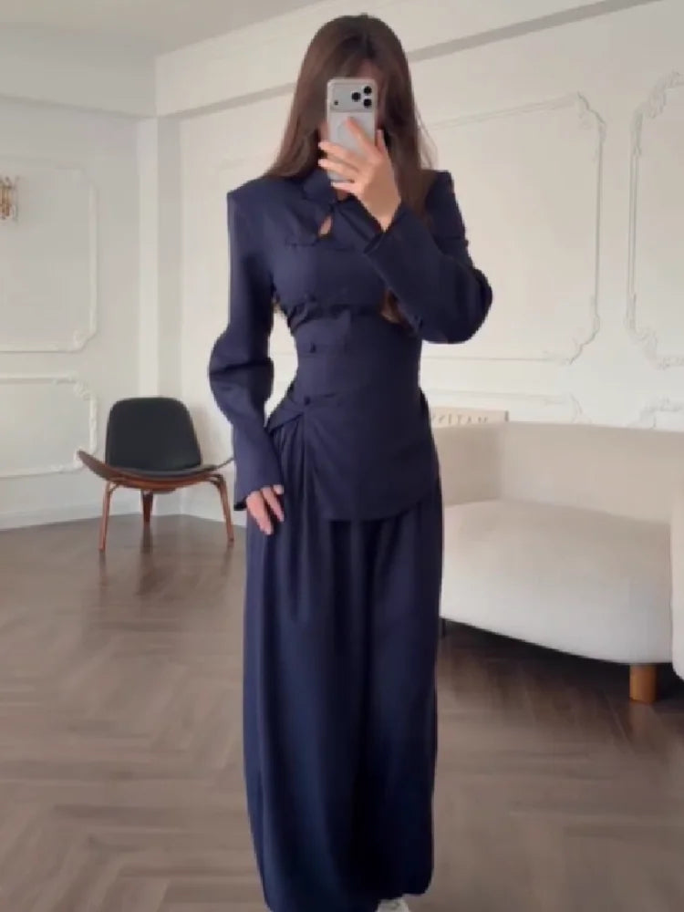 Person wearing a navy blue outfit taking a mirror selfie in a room with white walls and a chair.