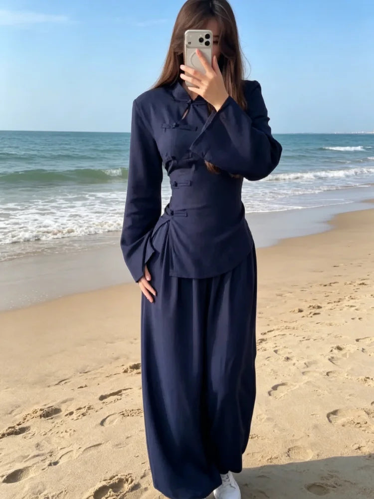 A woman takes a mirror selfie on a sandy beach wearing a dark navy blue traditional Chinese-style outfit consisting of a long-sleeve jacket with frog closures and matching wide-leg pants.