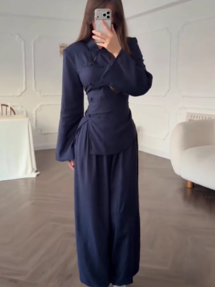 A woman takes a mirror selfie wearing a dark navy blue two-piece outfit consisting of a long-sleeve button-up top and matching wide-leg pants, in a bright room with white walls and wood floors.