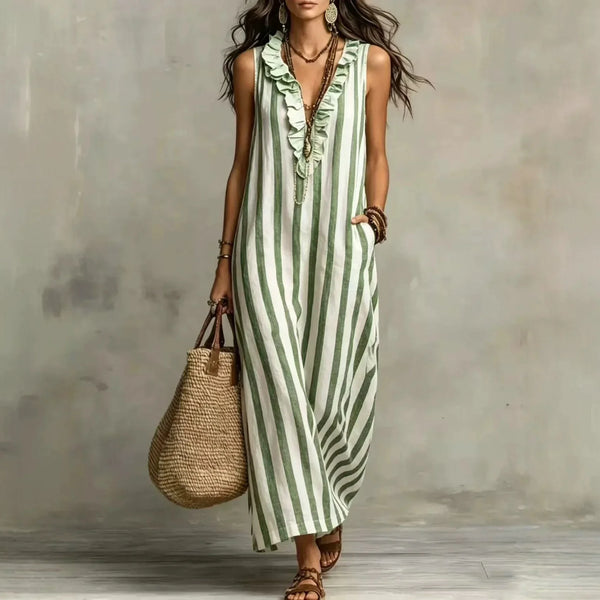 A woman models a sleeveless, ruffled V-neck maxi dress with vertical green and white stripes. She is holding a large woven straw tote bag and wearing gold jewelry, standing against a textured gray concrete wall.