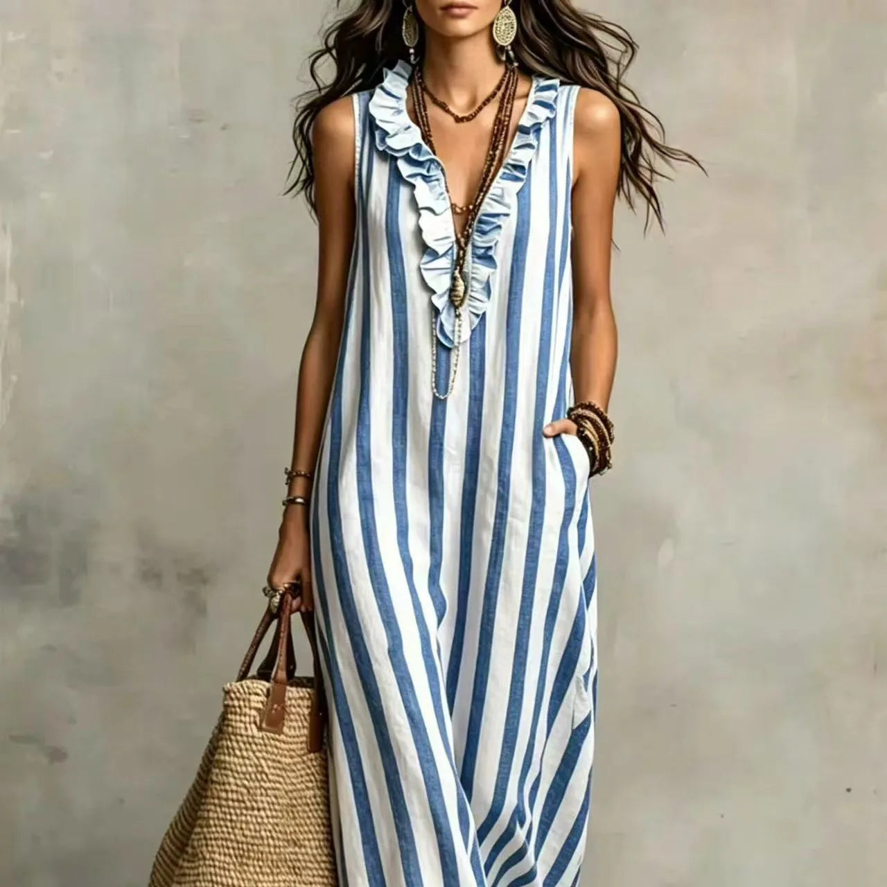 A woman wearing a sleeveless blue and white vertically striped maxi dress with a ruffled V-neckline, accessorized with layered necklaces, bracelets, and earrings, holding a woven straw tote bag against a textured gray wall.