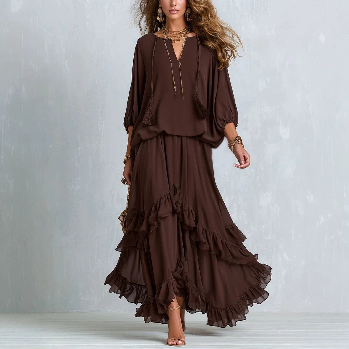A woman models a dark brown two-piece bohemian maxi dress set against a textured light gray studio backdrop. The set features a loose, flowy long-sleeve top with a deep V-neck and a matching tiered ruffle maxi skirt.