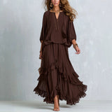 A woman models a dark brown two-piece bohemian maxi dress set against a textured light gray studio backdrop. The set features a loose, flowy long-sleeve top with a deep V-neck and a matching tiered ruffle maxi skirt.