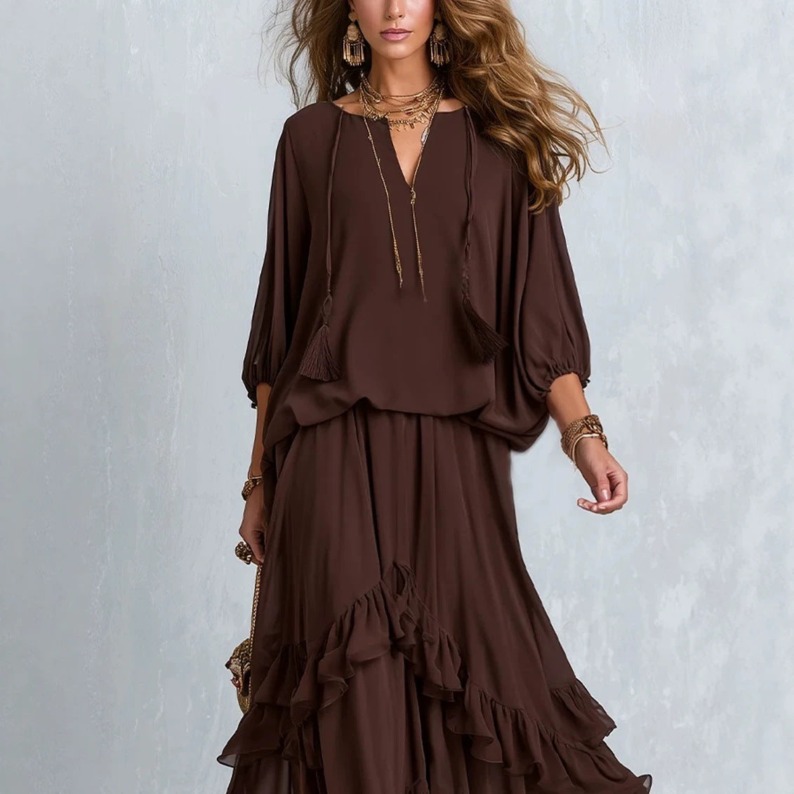 A woman models a dark brown two-piece outfit consisting of a long-sleeve blouse with drawstrings and a matching long skirt with ruffle details, set against a textured light gray background.