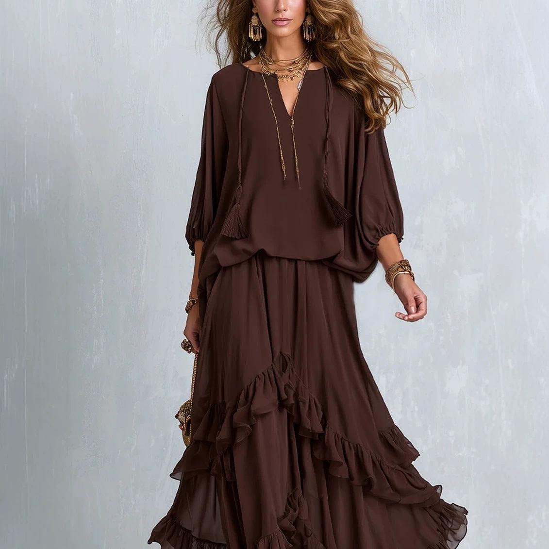 A woman models a dark brown two-piece outfit consisting of a loose long-sleeve top with a deep V-neck and a matching tiered maxi skirt, styled with gold jewelry against a textured gray background.