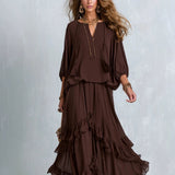 A woman models a dark brown two-piece outfit consisting of a loose long-sleeve top with a deep V-neck and a matching tiered maxi skirt, styled with gold jewelry against a textured gray background.
