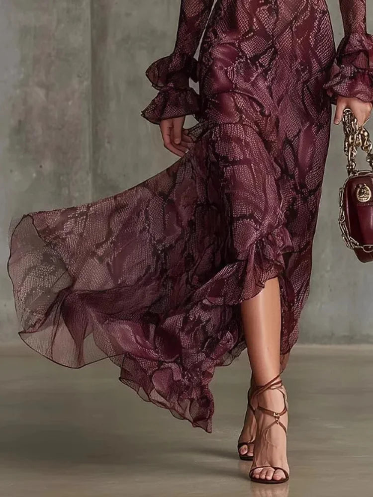A woman wearing a flowing burgundy snakeskin-print midi dress with ruffled sleeves and a side slit, paired with strappy high-heeled sandals and holding a matching burgundy handbag.