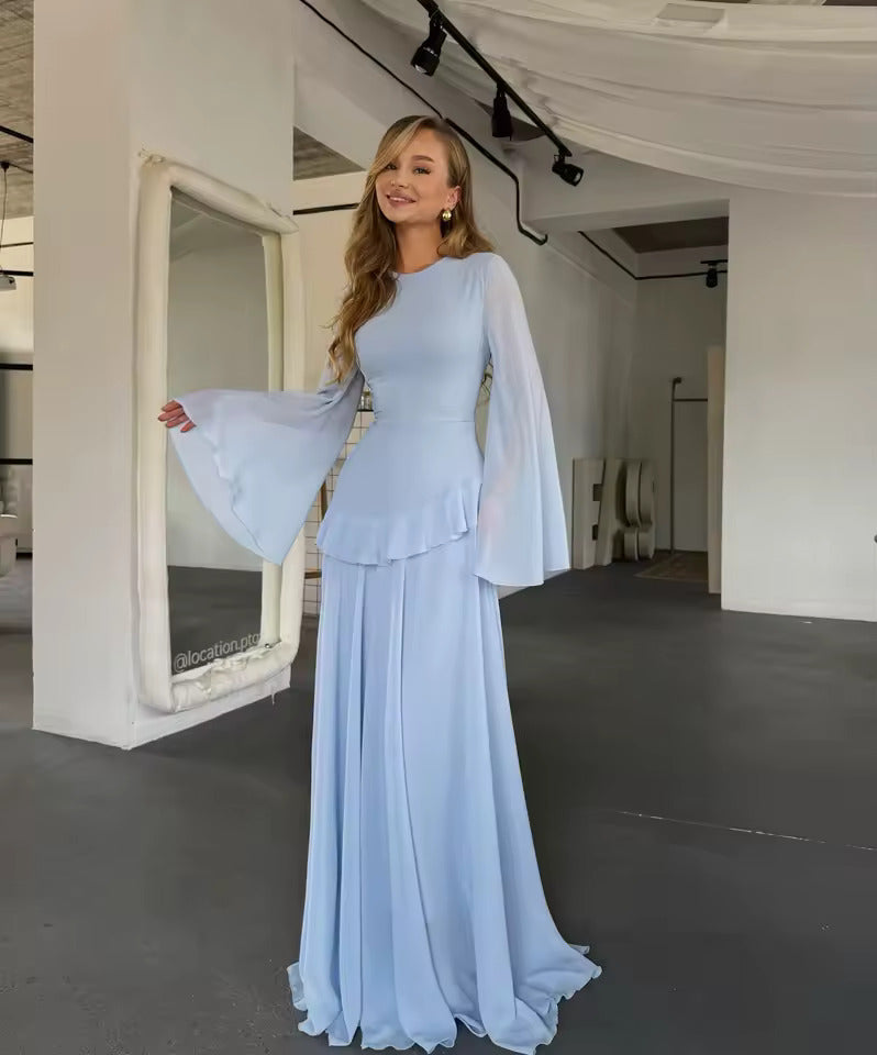 A woman models a light blue floor-length gown with a fitted bodice, ruffled waist, and dramatic sheer bell sleeves in a bright, modern studio with white walls and a large mirror.