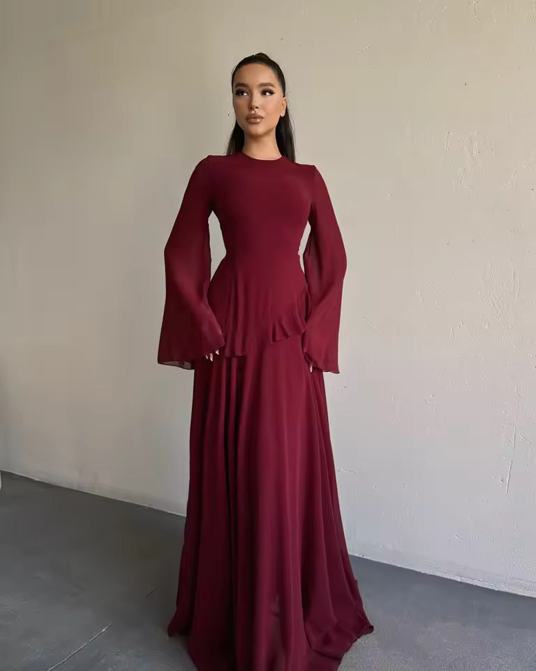 A woman wearing a long-sleeved, floor-length burgundy dress with a high neckline and flared bell sleeves, standing against a plain off-white wall.