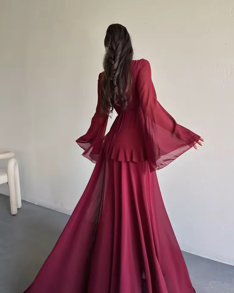 A woman viewed from behind wearing a long, flowing burgundy gown with dramatic flared sleeves and a layered ruffle waist, standing against a plain off-white wall.