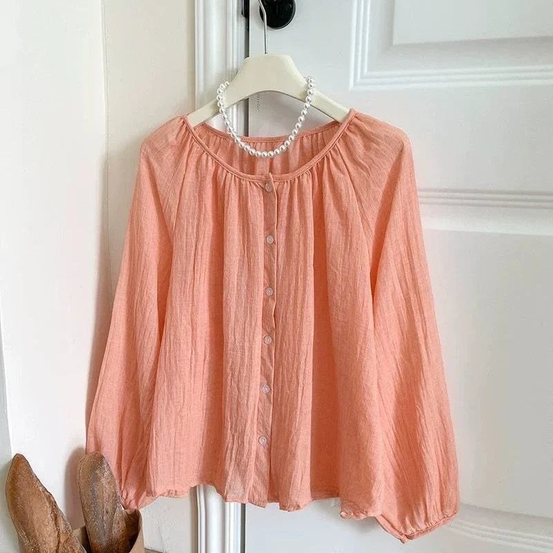 A peach-colored long-sleeve blouse with a button-down front, hanging on a white hanger decorated with a pearl necklace, set against a white paneled door.