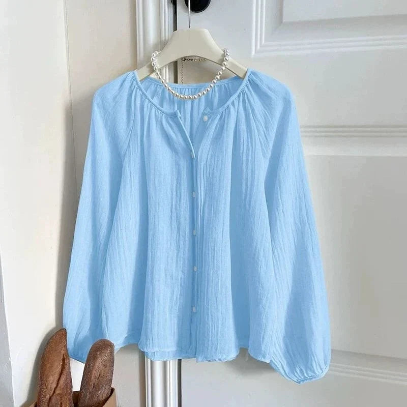 A light blue long-sleeve blouse with a gathered round neckline, button front, and slightly voluminous sleeves, hanging on a white hanger against a white paneled door. A pearl necklace is draped around the collar area.