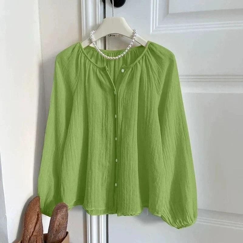 A light green crinkled blouse with a round neckline, button front, and long balloon sleeves, hanging on a white hanger against a white paneled door with a pearl necklace styled around the neck.