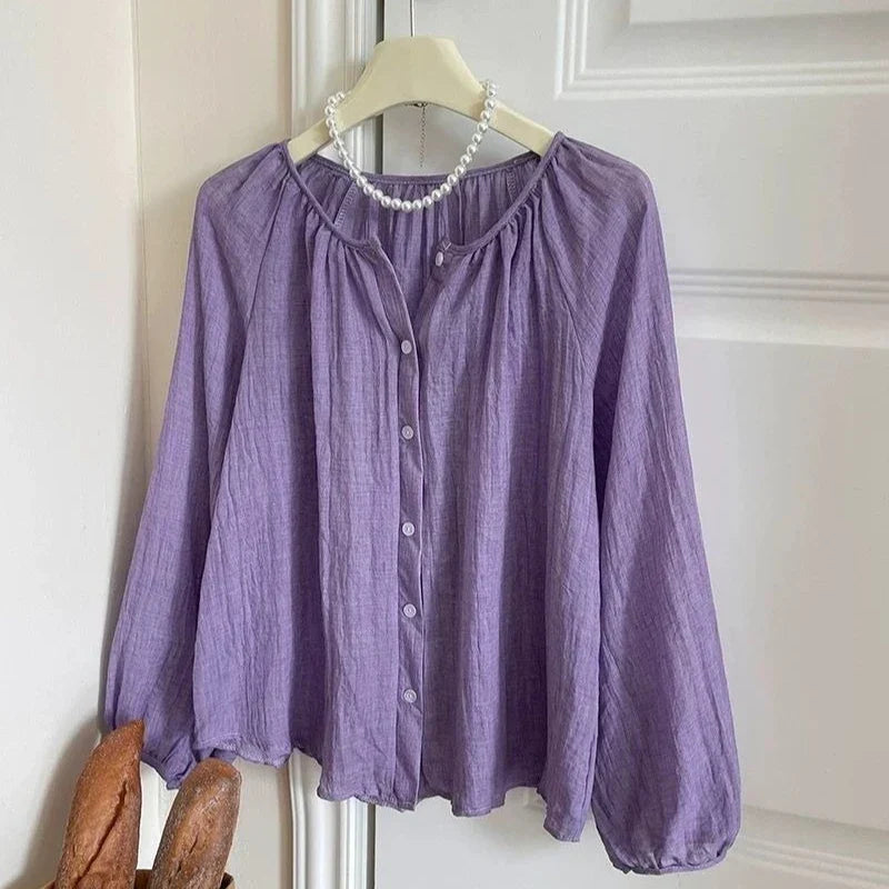 A lavender purple crinkle-textured blouse with a round neckline, button-up front, and gathered long sleeves, hanging on a cream hanger with a pearl necklace draped around the collar, set against a white paneled door.