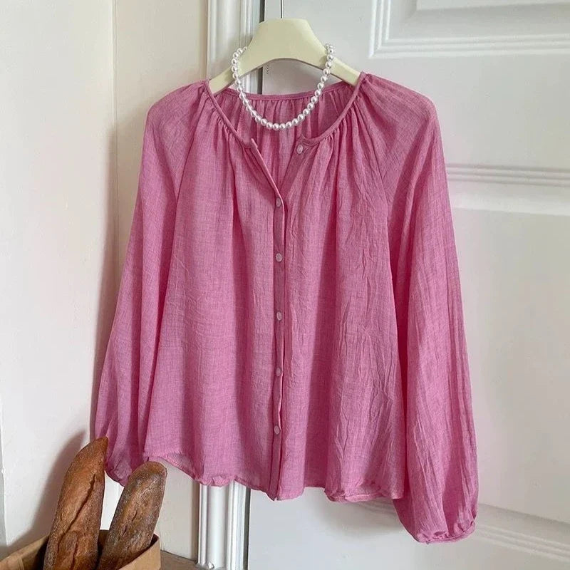 A pink long-sleeve linen blouse with a button-up front and gathered neckline, hanging on a white hanger decorated with a white pearl necklace. The blouse is styled with a basket of rustic bread loaves in the lower-left corner against a white paneled door.