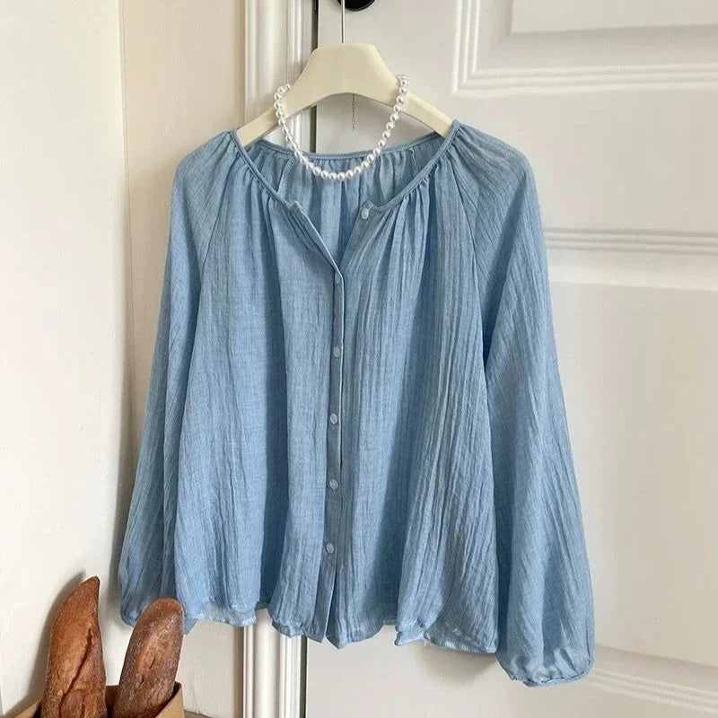 A light blue crinkled long-sleeve blouse hanging on a white hanger with a pearl necklace, set against a white paneled door background.
