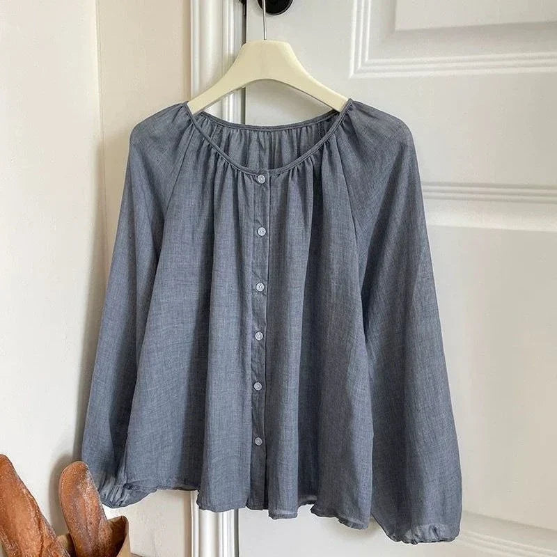 A slate blue-gray long-sleeve blouse with a gathered round neckline and a button-down front, hanging on a cream hanger against a white paneled door.