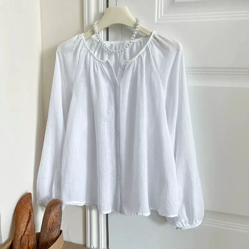 A white long-sleeve blouse with a gathered round neckline and button front, hanging on a hanger against a white paneled door. A pearl necklace is draped around the neck area, and a small decorative basket with bread handles is visible at the lower left.