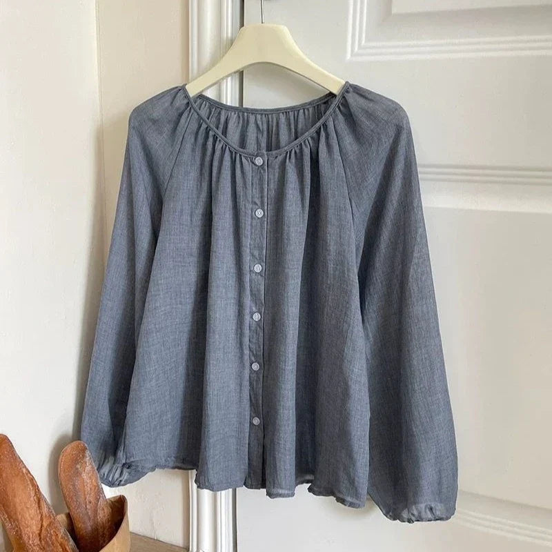 A slate blue-gray long-sleeve blouse with a gathered round neckline and a button-down front, hanging on a cream hanger against a white paneled door.