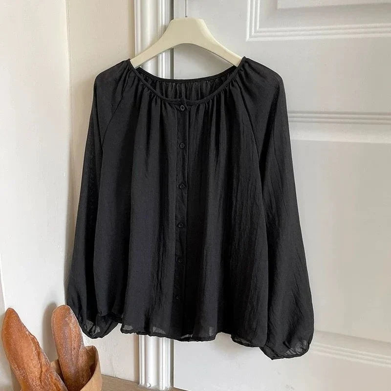 A black long-sleeve blouse with a gathered, elasticized neckline and a button-front closure, hanging on a white hanger against a white paneled door.