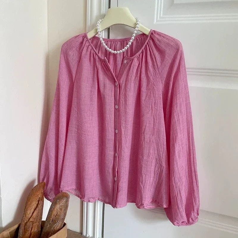 A pink long-sleeve blouse with a gathered round neckline, button-front closure, and slightly sheer textured fabric, hanging on a cream hanger against a white paneled door with a pearl necklace draped around the neck.