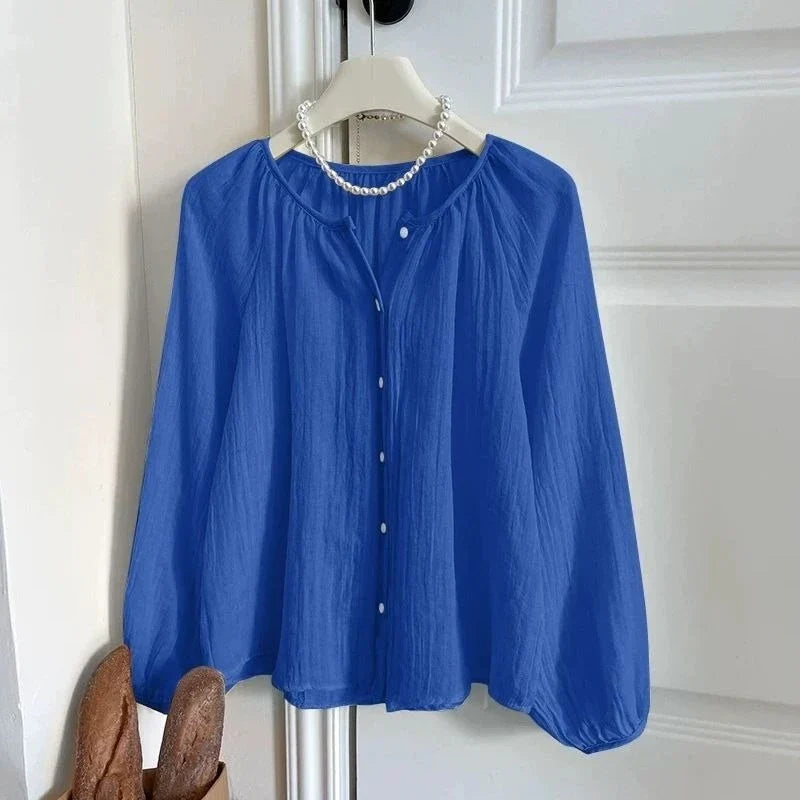 A royal blue long-sleeve blouse with a gathered round neckline, button-front closure, and voluminous sleeves, hanging on a white hanger against a white paneled door. A pearl necklace is draped around the neck area.