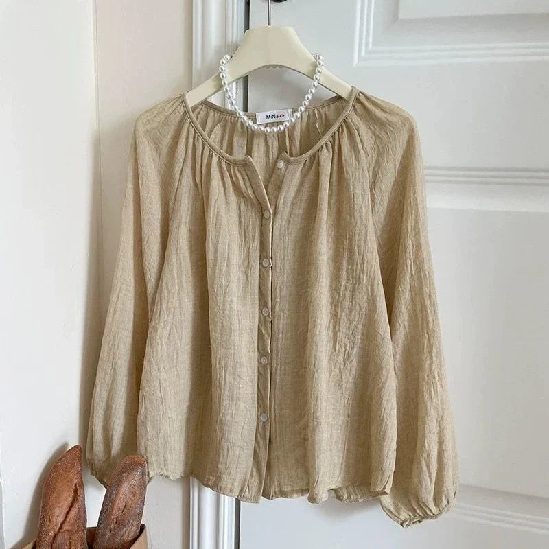 A beige linen blouse with a round neckline, button-up front, and gathered sleeves, hanging on a wooden hanger with a pearl necklace draped around the collar, set against a white paneled door.