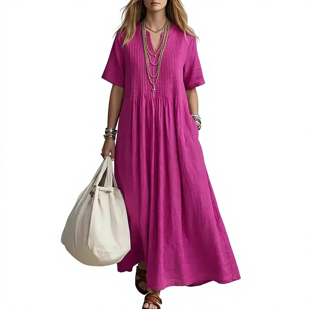 A woman wearing a bright magenta pink linen maxi dress with short sleeves, a gathered waist, and a pleated V-neckline. She is accessorized with layered necklaces, bracelets, and sandals, and is holding a large cream canvas tote bag against a plain white background.