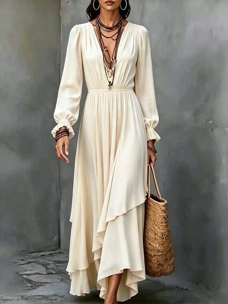 A woman models a long-sleeved cream-colored maxi dress with a deep V-neckline, gathered waist, and an asymmetrical draped hem. She is accessorized with layered necklaces, bracelets, and a woven straw tote bag, standing against a textured gray concrete wall.