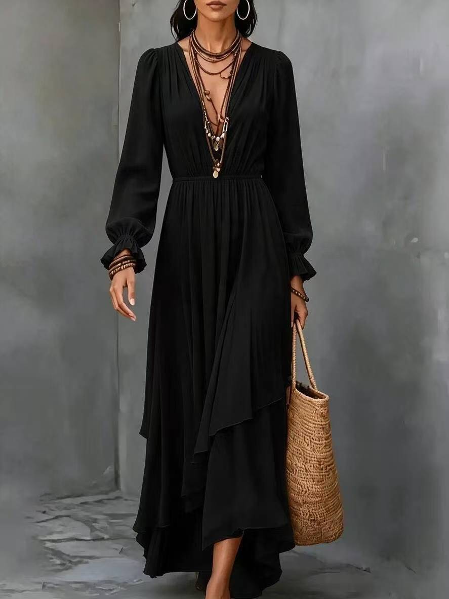 A woman models a long black dress with a deep V-neckline, long sleeves, and an asymmetrical draped hem. She is accessorized with layered necklaces, bracelets, and a woven straw tote bag, standing against a textured gray wall.