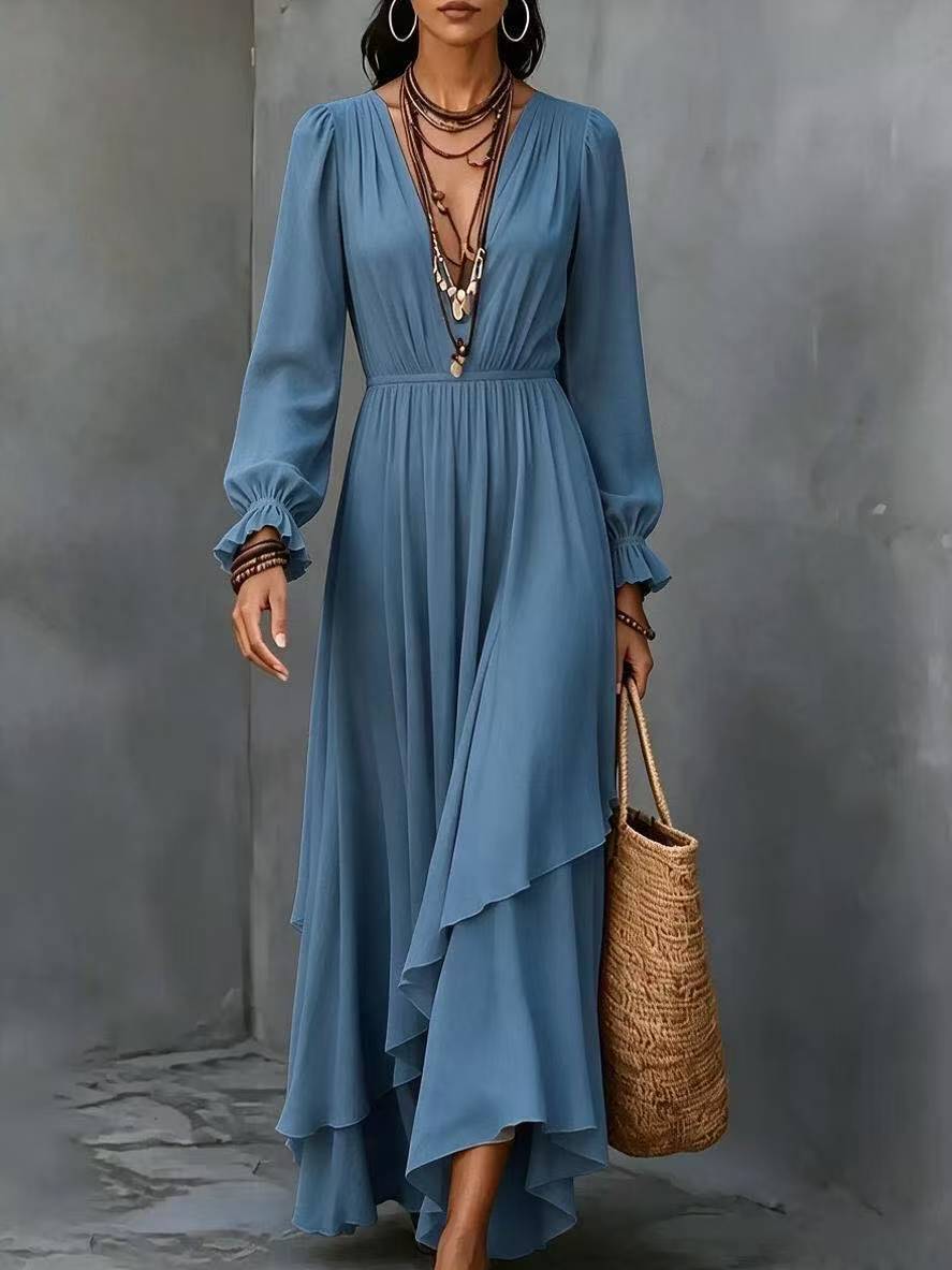 A woman wearing a dusty blue long-sleeve maxi dress with a gathered waist and layered ruffle hem, holding a woven straw tote bag against a textured gray wall.