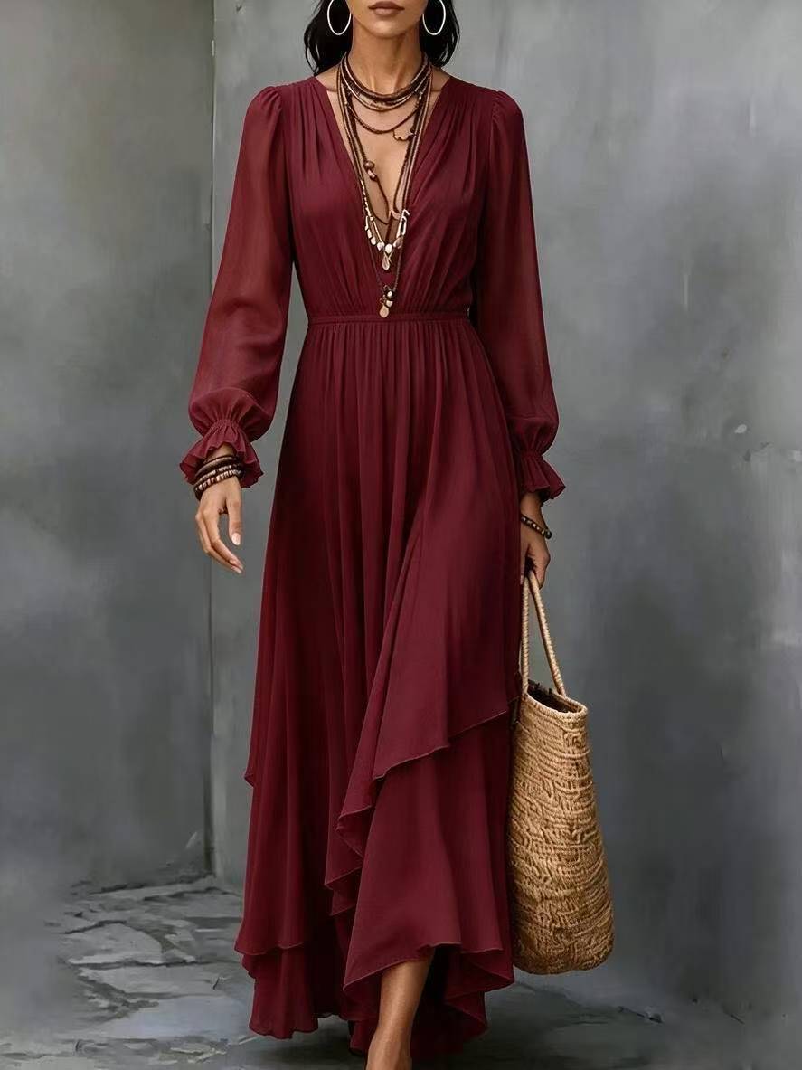 A woman models a long-sleeved, deep burgundy maxi dress with a high-low asymmetrical hem and layered necklaces, holding a woven straw tote bag against a textured gray wall.