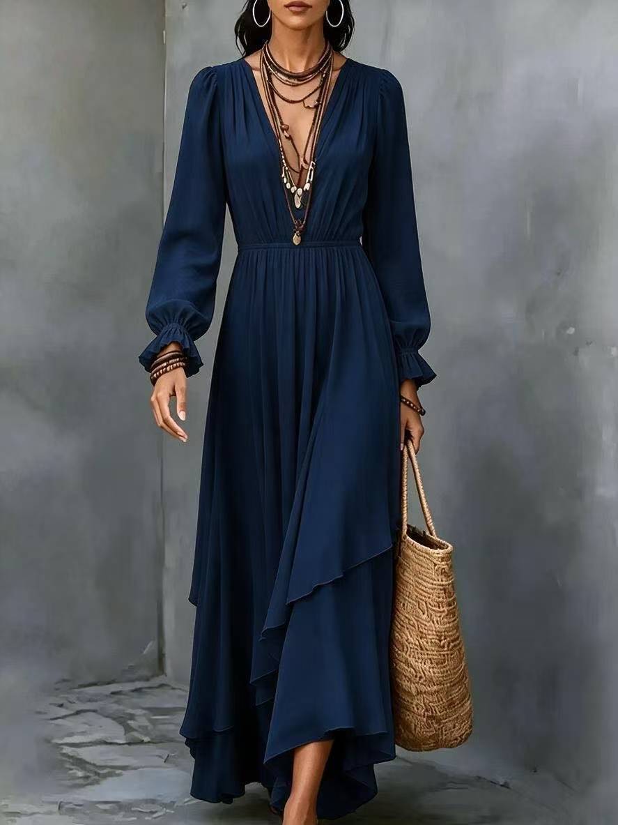 A woman models a long-sleeved navy blue maxi dress with a deep V-neckline, gathered waist, and layered asymmetrical ruffle hem. She is accessorized with layered necklaces, bracelets, hoop earrings, and carries a woven straw tote bag against a textured gray concrete wall.