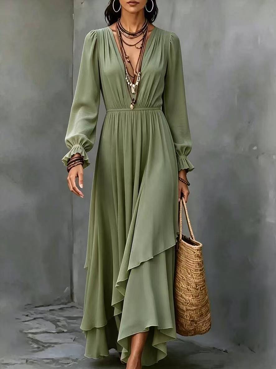 A woman models a sage green maxi dress with long sleeves, a deep V-neckline, and an asymmetrical layered hem. She is accessorized with layered necklaces, bracelets, and a woven straw tote bag, standing against a textured gray concrete wall.
