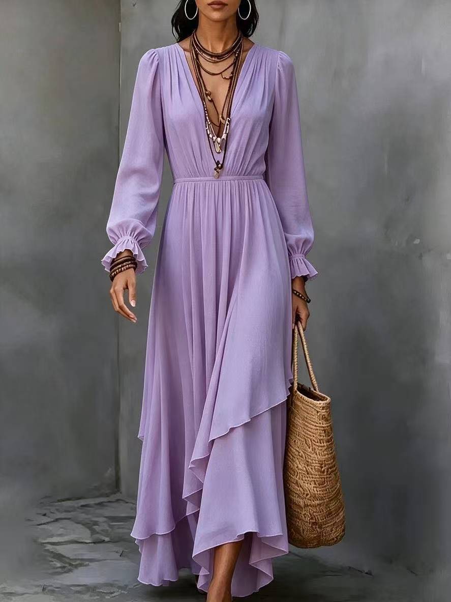 A woman models a long-sleeved lavender maxi dress with a high-low ruffled hem, holding a woven straw tote bag against a textured gray concrete wall.