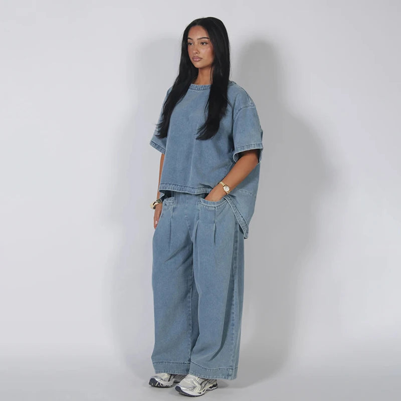A woman modeling a light blue denim two-piece set consisting of a short-sleeve cropped t-shirt and matching wide-leg pants, standing against a plain light gray studio background.