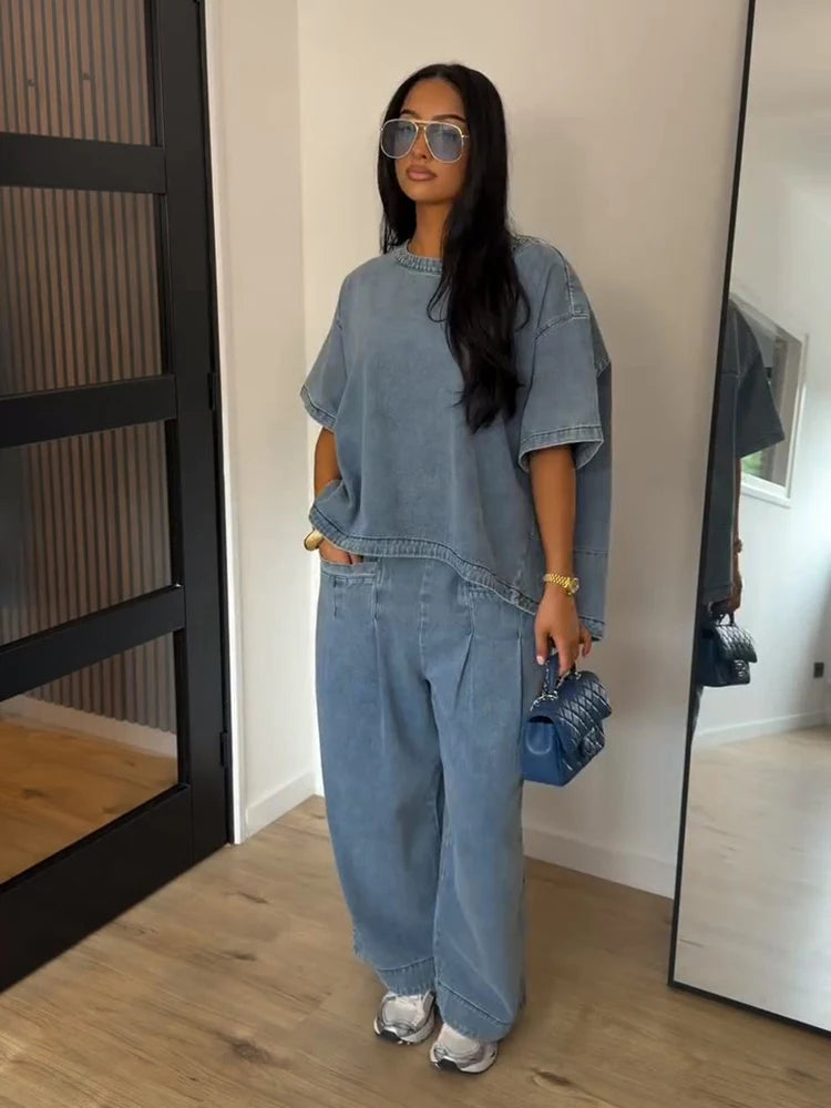 A woman stands indoors wearing an oversized light blue denim two-piece set with a short-sleeve top and wide-leg pants, paired with silver sneakers and holding a small blue handbag.