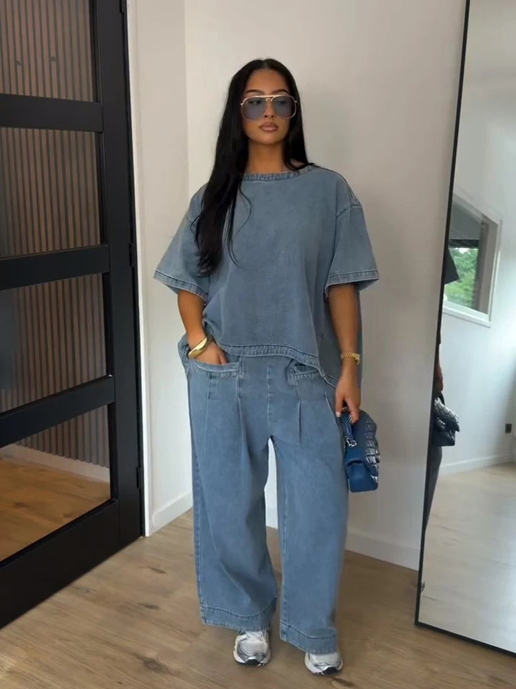 A woman modeling a matching light blue denim set consisting of a short-sleeve top and wide-leg pants, styled with sunglasses, a blue handbag, and white sneakers in an indoor setting.