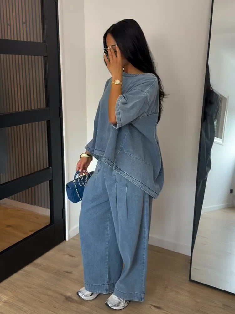A woman wearing a light blue denim two-piece outfit consisting of a loose short-sleeve top and matching wide-leg pants, standing in an indoor room with a mirror and wooden floor.