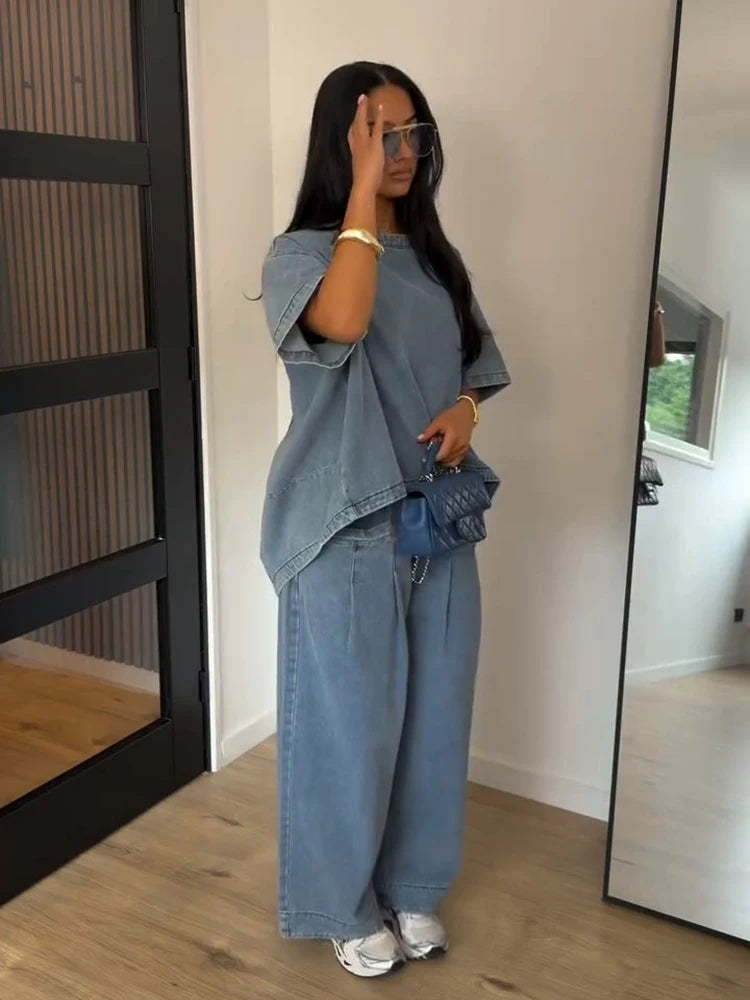A woman stands indoors wearing a light blue denim two-piece outfit consisting of a short-sleeve top and wide-leg pants, with a small blue quilted handbag and white sneakers.