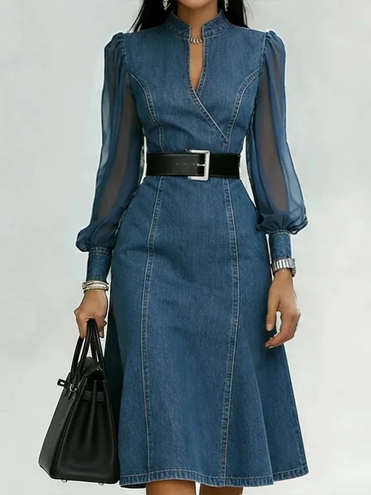 Blue denim dress with sheer sleeves and a black belt, held by a hand with a black bag on a white background.